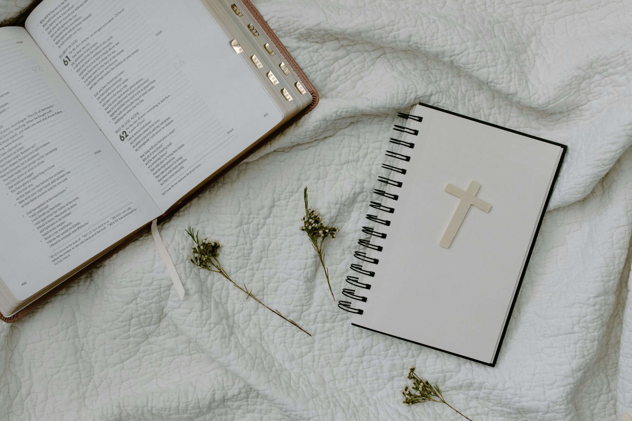 An open Bible and a cross-decorated notebook on white textured fabric with dried flowers.